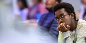 A student wearing glasses sits in a classroom while looking off screen with a focused look.