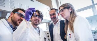 4 researchers, three in lab coats, one in a suit, are looking at a test tube together.